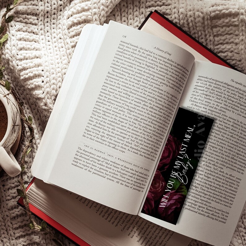 K&ouml;nnte beinhalten: Ein offenes Buch mit einem schwarzen Lesezeichen mit dem Text "WILL YOU BE MY LAST MEAL, Baby?" und roten Rosen. Eine Tasse Kaffee steht auf einer wei&szlig;en Strickdecke. Das Buch hat einen roten R&uuml;cken.