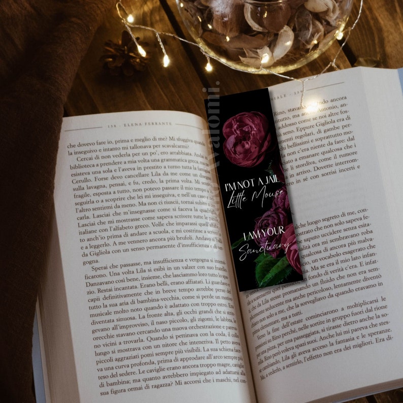 K&ouml;nnte beinhalten: Ein schwarzer Lesezeichen mit dem Text "I'm not a jail little mouse, I am your sanctuary" und Illustrationen von dunkelroten Rosen. Das Lesezeichen befindet sich in einem offenen Buch mit Text, mit Lichterketten und einer dekorativen Schale im Hintergrund.