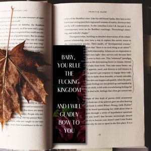K&ouml;nnte beinhalten: Ein schwarzer Lesezeichen mit wei&szlig;em Text, der lautet "BABY, YOU RULE THE FUCKING KINGDOM AND I WILL GLADLY BOW TO YOU", liegt auf einem offenen Buch mit einem getrockneten Blatt. Das Lesezeichen hat ein Blumendesign.
