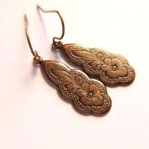 May include: A pair of bronze-coloured earrings with a floral design. The earrings have an oval shape with an embossed flower and leaf pattern. They feature a hook closure. The earrings are made of metal.