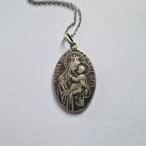 Op de afbeelding: Zilveren ketting met hanger met een religieus beeld van de Maagd Maria die het kindje Jezus vasthoudt. De hanger is ovaalvormig en heeft de inscriptie "Regina Deo Carmel O.P.N."