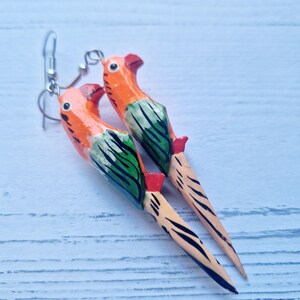 Op de afbeelding: Een paar houten papegaaioorbellen met oranje, groene en zwarte verf. De oorbellen hebben zilveren haakjes.