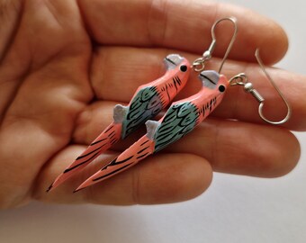 May include: A pair of parrot-shaped earrings. The earrings are hand-painted with pink, blue, and black details. The parrot's head and body are pink, with blue wings and black accents. The earrings have silver-colored hooks.