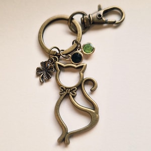 May include: Antique bronze cat-shaped keychain with a bow tie, a four-leaf clover charm, and two green gemstone accents. The keychain includes a clasp and a key ring.
