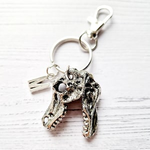 May include: Silver dinosaur skull keychain with a silver letter M charm attached. The keychain is on a white wooden surface.