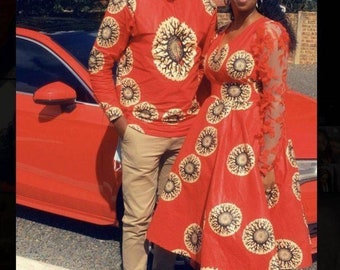 May include: A couple wearing matching red outfits with a sunflower pattern. The man is wearing a long-sleeved shirt and khaki pants. The woman is wearing a dress with lace sleeves. They are standing in front of a red car.