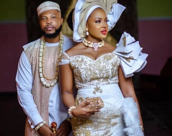 May include: A couple in traditional Nigerian wedding attire. The woman is wearing a white and gold embroidered dress with a large white head wrap. The man is wearing a white shirt, a long white robe, and a beaded necklace.