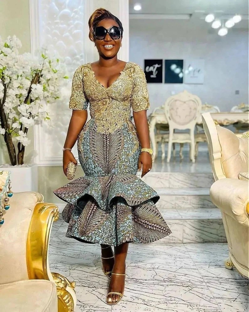 Pode incluir: Uma mulher vestindo um vestido estampado em dourado e azul com uma saia rodada. Ela est&aacute; usando &oacute;culos de sol e segurando uma pequena carteira.