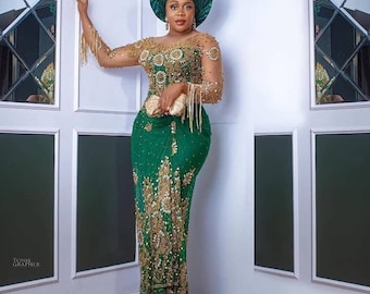 Pode incluir: Uma mulher vestindo um vestido longo verde com bordados dourados e um turbante verde combinando. O vestido tem um corpete justo e uma saia fluida. A mulher está em pé em um quarto branco com um piso de madeira.