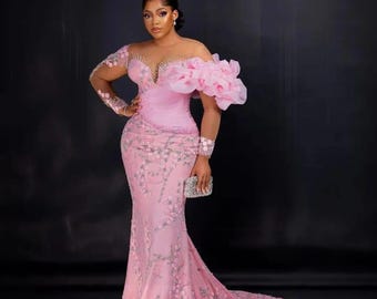May include: A pink, long-sleeved, mermaid-style gown with a sheer bodice and a large, ruffled flower on one shoulder. The gown is decorated with delicate floral embroidery.