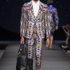 May include: A man wearing a patterned suit in shades of red, blue, and green. He is wearing sunglasses and carrying a black leather bag.