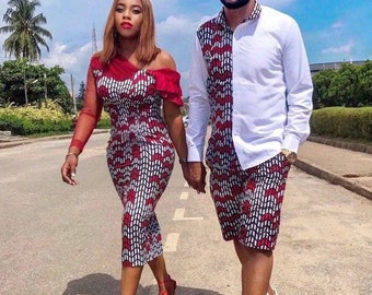 May include: A couple wearing matching red, white, and black patterned outfits. The woman is wearing a dress with a one-shoulder design and the man is wearing a shirt and shorts. They are walking down a street together.
