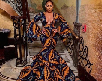 May include: A woman wearing a long, fitted dress with a plunging neckline and bell sleeves. The dress is made of a colorful fabric with a geometric pattern. The woman is standing on a staircase with a decorative railing.