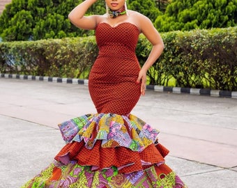 May include: A woman wearing a red and patterned dress with a fitted bodice and a large, multi-layered skirt. She is wearing a colourful head wrap and a necklace.