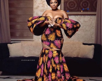 May include: A woman wearing a black, pink, and gold patterned dress with a large, dramatic skirt. The dress has a sweetheart neckline and large, puffy sleeves. The woman is standing in a room with a couch and a chandelier in the background.