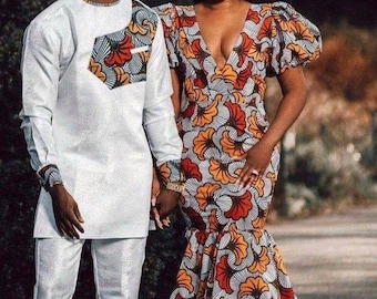 May include: A couple wearing matching traditional African clothing. The man is wearing a white long-sleeved shirt with a patterned chest panel and white pants. The woman is wearing a long, fitted dress with a V-neckline and a colorful floral pattern.