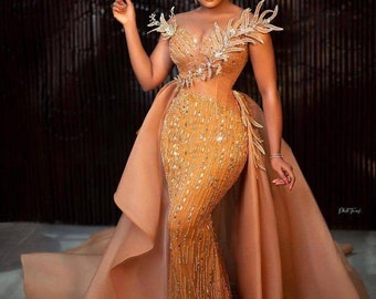 May include: A woman wearing a long, flowing, gold and brown sequined gown with a high neckline and a train. The gown has a fitted bodice and a mermaid skirt. The woman is standing in front of a dark background.