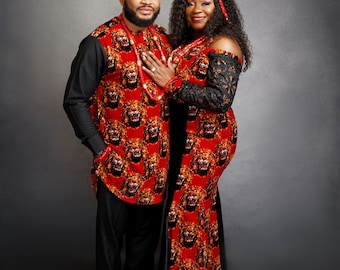 May include: A couple in matching red and black outfits. The man wears a long-sleeved shirt and pants, and a cap, all with a lion face pattern. The woman wears a matching dress with black lace sleeves and a red headband. Both wear beaded necklaces.