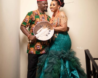 May include: A couple in traditional Nigerian wedding attire. The woman is wearing a teal green gown with a ruffled skirt and a beaded headpiece. The man is wearing a green shirt with a lion print and a red fez. They are holding a sign that says "Awoma Ig"