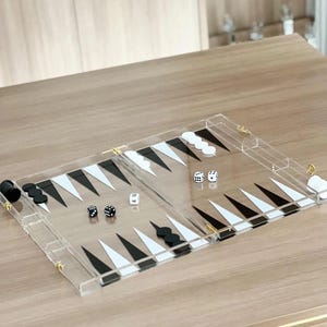 May include: A clear acrylic backgammon set with black and white triangular points. The set includes black and white game pieces and dice. The board is on a light brown wooden surface.