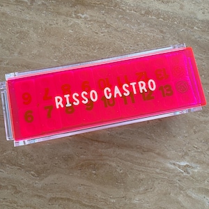 May include: A rectangular, clear acrylic box with a bright pink interior. The box contains numbered cards and the text "RISSO CASTRO" in white. The numbers are gold and range from 6 to 13. The box is resting on a light brown surface.