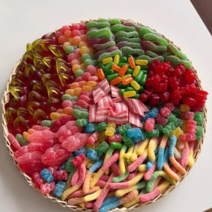 Könnte beinhalten: Ein runder Weidenkorb gefüllt mit einer bunten Auswahl an Gummibonbons. Die Süßigkeiten umfassen Gummibärchen, Würmer und andere Formen in verschiedenen Farben wie Rot, Grün, Gelb und Blau. Der Korb steht auf einer weißen Oberfläche.