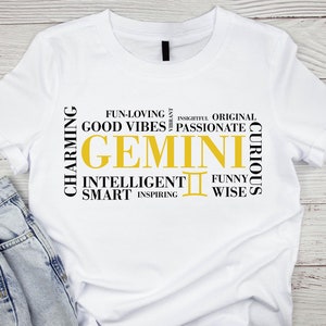 May include: White t-shirt with a black and gold graphic design featuring the word "GEMINI" surrounded by words describing Gemini traits, such as "charming", "intelligent", "funny", and "inspiring".
