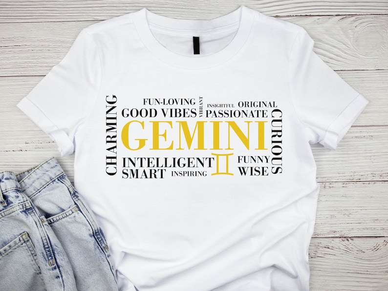 May include: White t-shirt with a black and gold graphic design featuring the word "GEMINI" surrounded by words describing Gemini traits, such as "charming", "intelligent", "funny", and "inspiring".
