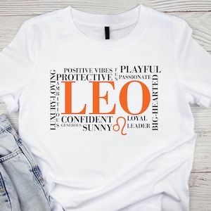 May include: White t-shirt with a black and orange graphic design featuring the word "LEO" surrounded by words describing Leo personality traits, such as "protective", "passionate", "confident", "loyal", and "leader".