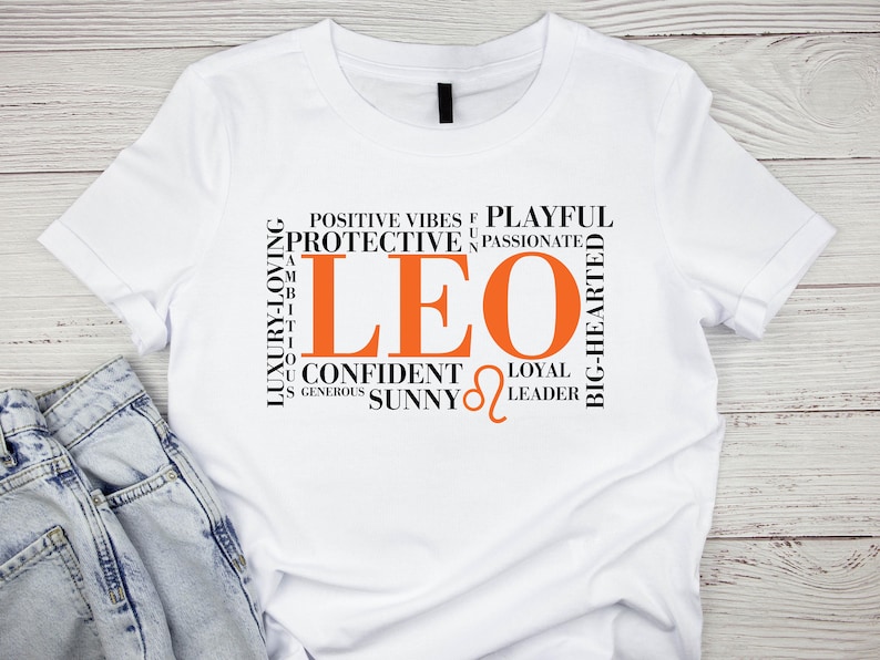 May include: White t-shirt with a black and orange graphic design featuring the word "LEO" surrounded by words describing Leo personality traits, such as "protective", "passionate", "confident", "loyal", and "leader".