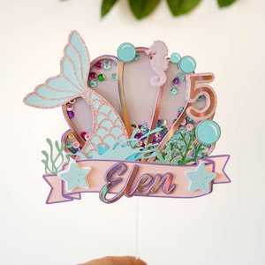 Op de afbeelding: Een caketopper met een zeemeermin-thema met een roze en blauwe schelp, een zeemeerminstaart, een zeepaardje en de naam "Elen" op een banner. De topper is versierd met glitter en pailletten.