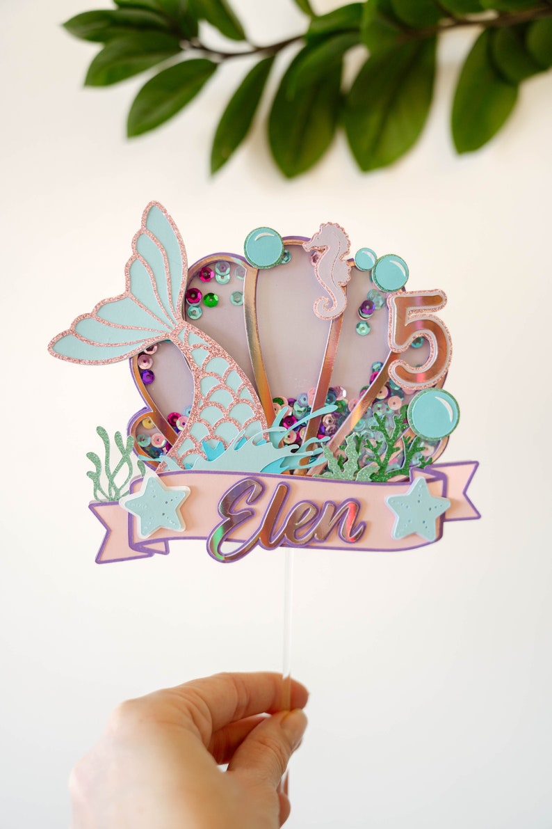 Op de afbeelding: Een caketopper met een zeemeermin-thema met een roze en blauwe schelp, een zeemeerminstaart, een zeepaardje en de naam "Elen" op een banner. De topper is versierd met glitter en pailletten.