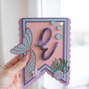 May include: A pink and purple banner featuring a mermaid tail, the letter "E", and sea life decorations. The banner is decorated with glitter and has a scalloped edge.