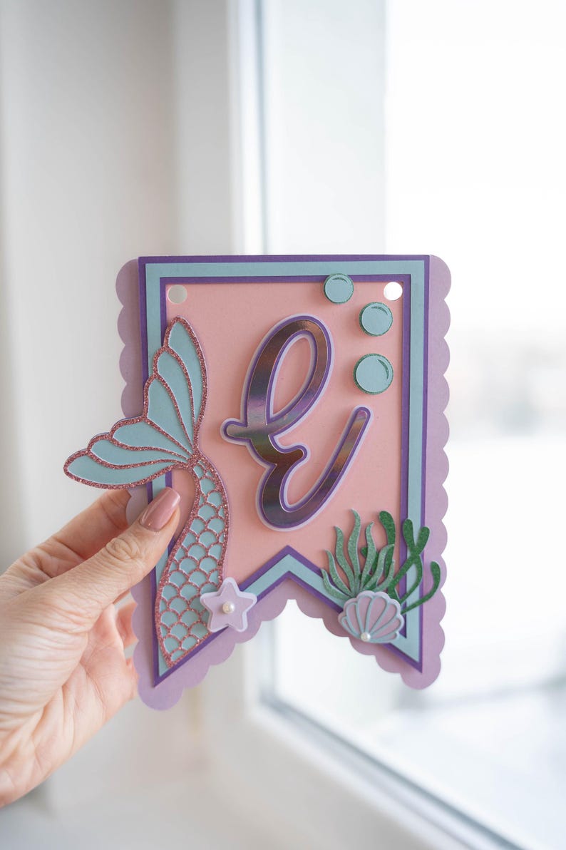 May include: A pink and purple banner featuring a mermaid tail, the letter "E", and sea life decorations. The banner is decorated with glitter and has a scalloped edge.