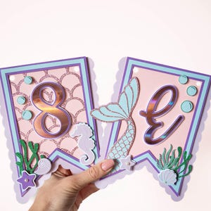 May include: A pink and purple mermaid-themed banner with the number "8" and the letter "E" in gold foil. The banner features a mermaid tail, seashells, and seaweed.
