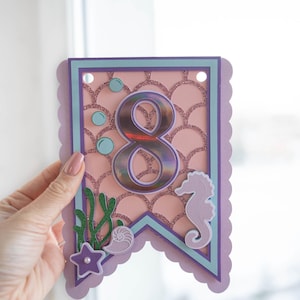 May include: Purple and pink mermaid-themed banner featuring a large iridescent number 8, a seahorse, a seashell, and seaweed. The banner has a scalloped edge and is decorated with glitter.