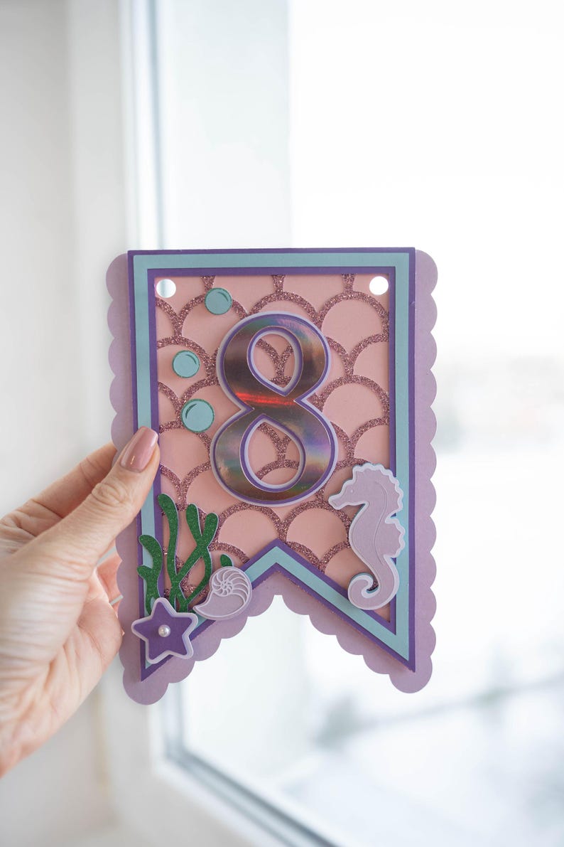 May include: Purple and pink mermaid-themed banner featuring a large iridescent number 8, a seahorse, a seashell, and seaweed. The banner has a scalloped edge and is decorated with glitter.