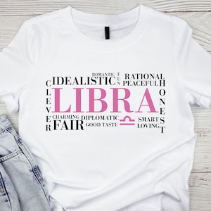 May include: White t-shirt with a pink Libra zodiac sign and a word cloud of traits associated with the sign, including "idealistic", "charming", "diplomatic", "fair", "romantic", "rational", "peaceful", "smart", "loving", and "good taste".