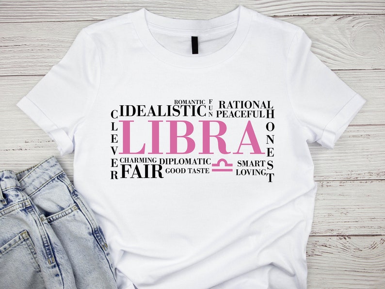May include: White t-shirt with a pink Libra zodiac sign and a word cloud of traits associated with the sign, including "idealistic", "charming", "diplomatic", "fair", "romantic", "rational", "peaceful", "smart", "loving", and "good taste".