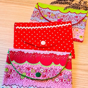 May include: Three fabric pouches with different patterns. The top pouch is light blue with green trim and a white button. The middle pouch is red with white polka dots and a white button. The bottom pouch is pink with a green trim and a green button.