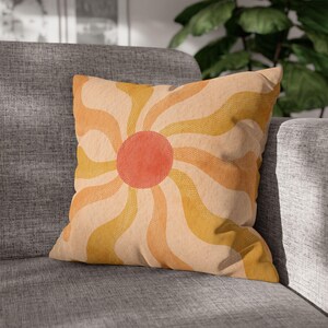 May include: Square decorative pillow with a retro sunburst design. The pillow features a central orange circle with radiating wavy lines in shades of yellow and orange against a beige background. The pillow is placed on a gray sofa.