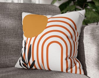 May include: A white throw pillow with an orange and black abstract design. The design features a large orange circle, a black leaf, and a series of orange stripes forming an arch.