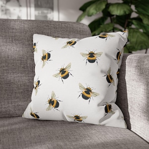 May include: Square white throw pillow with a repeating pattern of black and yellow bumblebees. The pillow is placed on a gray sofa. The bees are depicted with detailed wings and body markings, creating a nature-inspired design.