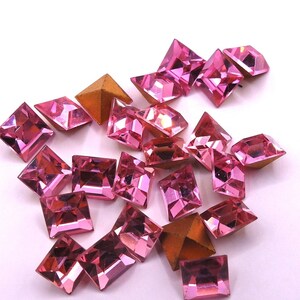 May include: A collection of pink, faceted gemstones in various shapes, including squares, rectangles, and triangles. The gems have a reflective surface and are scattered on a white background. Some gems have a gold-colored backing.