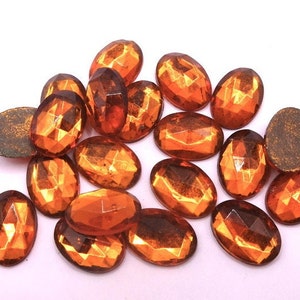 Puede incluir: Un conjunto de gemas ovales y facetadas de color naranja. Las gemas tienen una superficie reflectante y un reverso plano y texturizado. Las piedras están dispersas sobre un fondo blanco, resaltando su color y corte.