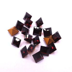 May include: A scattering of square-cut, deep red gemstones and two golden pyramid-shaped objects on a white background. The gemstones have a faceted appearance, reflecting light. The arrangement suggests a focus on jewellery or crafting materials.