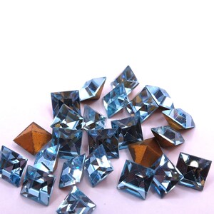May include: A selection of light blue, square-cut gemstones with various facets. Some gemstones have a metallic, bronze-coloured backing. The gems are scattered against a white background, creating a sparkling effect. Suitable for jewellery making.