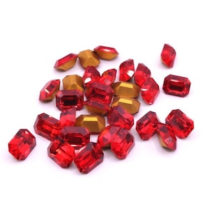 May include: A collection of red and gold-coloured faceted gemstones. The red gems have a rectangular emerald cut, while the gold gems have a more geometric shape. The gems are scattered on a white surface.