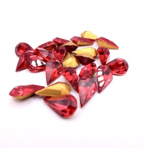 May include: A selection of red, teardrop-shaped rhinestones with gold-coloured backs. The rhinestones are scattered on a white surface, with varying sizes and facets that catch the light. Suitable for craft and embellishment projects.