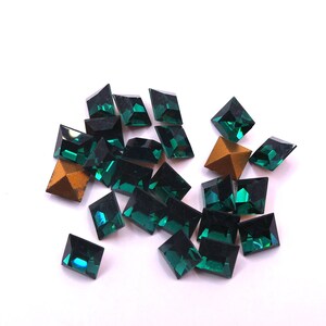 May include: A collection of emerald green glass rhinestones in various geometric shapes, including squares and trapezoids. Some have a metallic gold finish. The gemstones are scattered on a white surface.